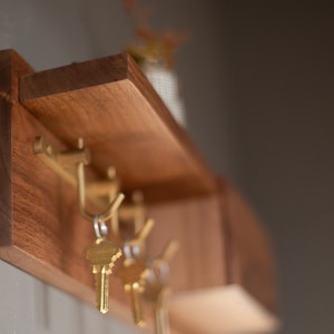 May include: A wooden wall-mounted key rack with a shelf. The rack has four hooks and is made of dark wood. There are keys hanging on the hooks.