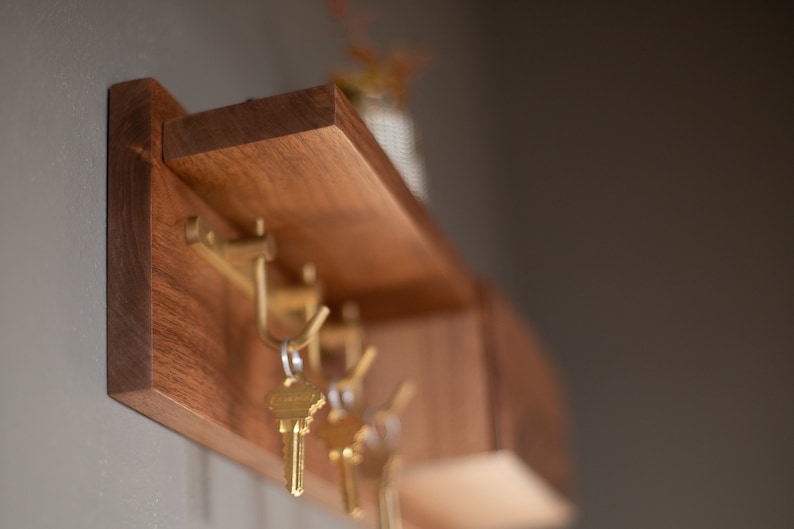 May include: A wooden wall-mounted key rack with a shelf. The rack has four hooks and is made of dark wood. There are keys hanging on the hooks.