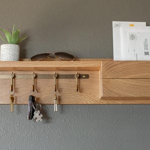 May include: A wooden wall-mounted key holder with a shelf and a mail slot. The key holder has five hooks and a brass bar. There are keys and sunglasses on the hooks. A small potted plant sits on the shelf.