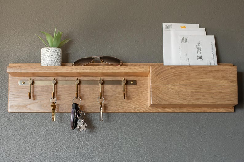 May include: A wooden wall-mounted key holder with a shelf and a mail slot. The key holder has five hooks and a brass bar. There are keys and sunglasses on the hooks. A small potted plant sits on the shelf.