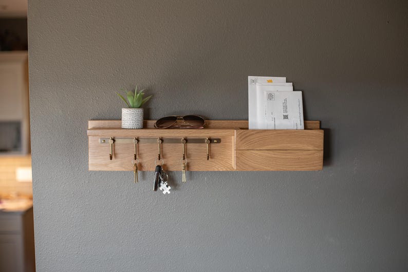 May include: A wooden wall-mounted key holder with a built-in mail slot. The key holder has five hooks and a small shelf for decorative items. There is a potted succulent plant and a pair of sunglasses on the shelf. There are three envelopes in the mail slot.