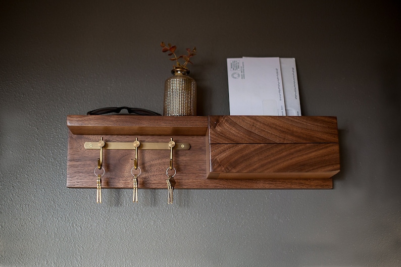 May include: A wooden wall mounted key holder with a shelf and a mail slot. The key holder has four brass hooks and a brass bar. There are three keys hanging on the hooks. A small vase with a plant is on the shelf. There are two white envelopes in the mail slot.