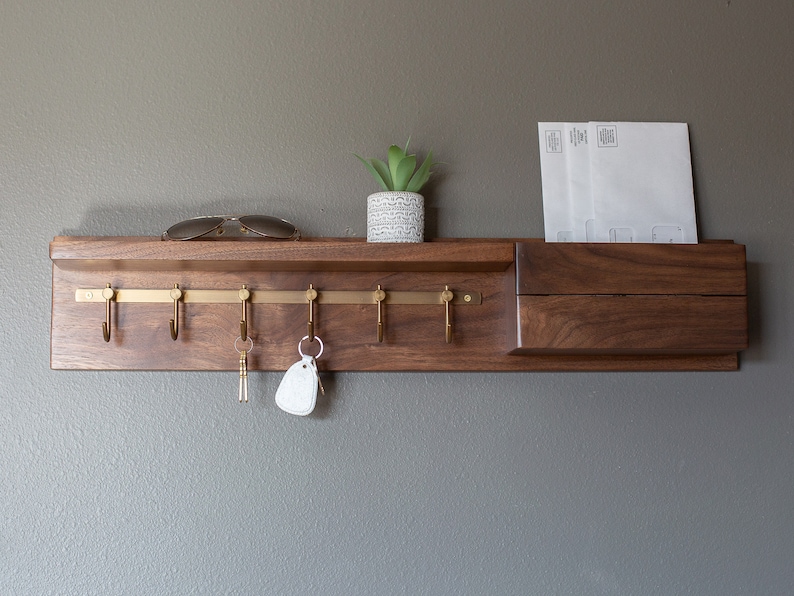 May include: A wooden wall-mounted key rack with a shelf and a mail slot. The rack has six brass hooks and a small potted plant on the shelf.