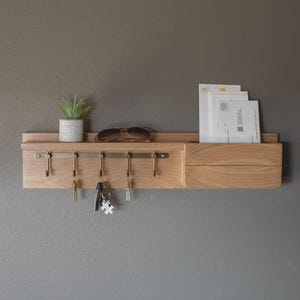 May include: A wall-mounted light wood organizer featuring a horizontal shelf, five metal hooks for keys, and a side compartment for mail. The shelf holds a small potted succulent and sunglasses, while the side slot contains white envelopes.