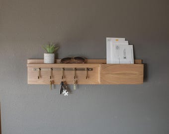 May include: A wall-mounted light wood organizer featuring a horizontal shelf, five metal hooks for keys, and a side compartment for mail. The shelf holds a small potted succulent and sunglasses, while the side slot contains white envelopes.