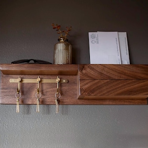 May include: A wall-mounted dark wood entryway organizer featuring a horizontal shelf, a brass metal bar with three hooks holding keys, and a small storage compartment. A textured gold vase with dried foliage and white envelopes sit on the shelf.