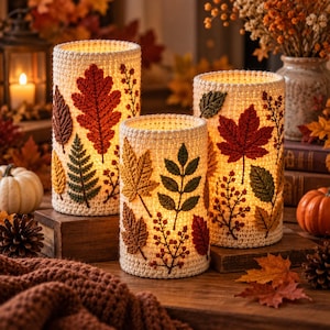 May include: Three cylindrical crochet candle covers in cream yarn, each decorated with appliqued autumn leaves in shades of red, orange, yellow, and green. The covers are illuminated from within, highlighting the textured stitch patterns.