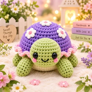 Puede incluir: Un peluche de tortuga tejido a ganchillo con cabeza y cuerpo verde claro, con un caparazón morado decorado con flores blancas y un borde rosa con volantes. La tortuga tiene grandes ojos negros, una sonrisa bordada y mejillas rosadas.