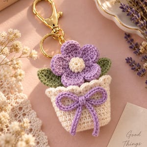 May include: A crochet keyring featuring a small cream-coloured pot with a purple bow, containing a purple flower with a cream centre and two green leaves. The item is attached to a gold-toned metal lobster clasp and a circular keyring for attachment.