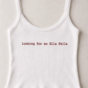 Op de afbeelding: Een wit geribbelde tanktop met dunne schouderbandjes en een ronde halslijn. In het midden van de borst staat de tekst "looking for an Ella Fella" geborduurd in een donkerrood, kleine letters serif lettertype. De zoom heeft een subtiele golvende rand.