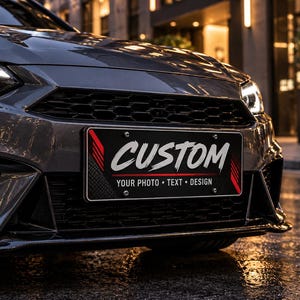 May include: A rectangular black license plate mounted on the front bumper of a dark gray car. The plate features the white text CUSTOM in a stylized font, with red graphic accents on the sides and the text YOUR PHOTO • TEXT • DESIGN below.
