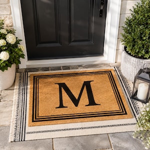 May include: A rectangular tan coir doormat featuring a large black letter M in the center, surrounded by a triple-line black border. The mat is layered on top of a larger, off-white rectangular rug with black striped accents and fringed edges.