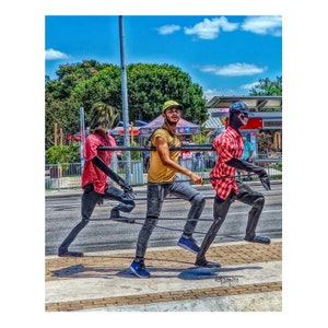 May include: Three people dressed in black and white clothing are walking in a street scene. The person in the middle is wearing a yellow shirt and jeans. The person on the left is wearing a red shirt and the person on the right is wearing a red and white plaid shirt.