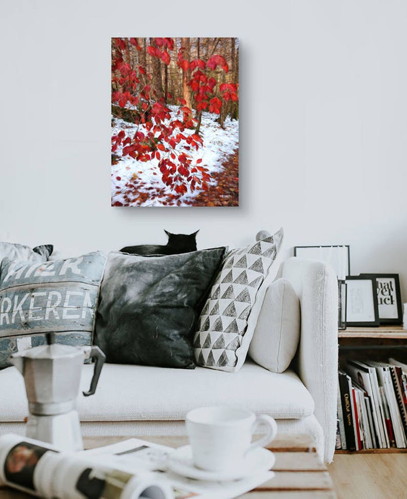 May include: A framed canvas print depicting a forest scene with striking red leaves set against snow and trees. The artwork is displayed above a white sofa with cushions, a coffee maker, and a cup.
