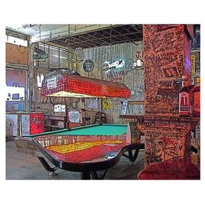 May include: A green felt pool table with a red wooden frame in a bar setting. The table is lit by a red and white checkered light fixture. The wall behind the table is covered in graffiti and writing. A Budweiser sign is visible on the left side of the image.