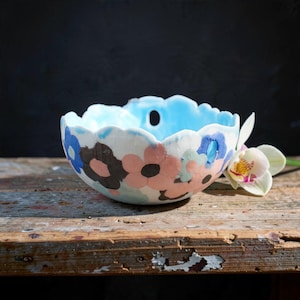 May include: A ceramic bowl with a scalloped rim and a light blue interior. The exterior features a pattern of large, flat-coloured flowers in shades of pink, dark brown, and blue. A single small hole is visible near the top rim of the bowl.