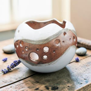 May include: A spherical ceramic bowl with a wavy rim, featuring a smooth white glaze on the top and bottom sections and a textured, metallic copper-toned band around the center, punctuated by several circular cutouts of varying sizes.