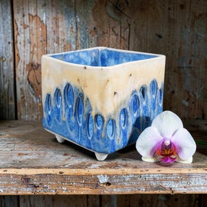 May include: A square ceramic planter with a cream-colored top and a blue base featuring vertical cutouts that create a dripping effect. The planter stands on four small feet and is positioned next to a white orchid flower with a purple center.