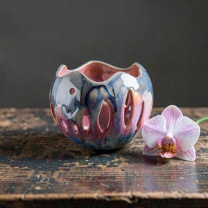 May include: A spherical ceramic candle holder with a wavy top edge and vertical cutouts. The glaze features a blend of deep blue, purple, and pink tones. A single light pink orchid flower with a yellow and dark pink center sits beside it.
