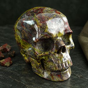 May include: A polished stone skull carving featuring a mottled pattern of deep olive green, dark forest green, and rich burgundy red. The surface is smooth and reflective, showcasing the natural mineral variations in a realistic human skull shape.