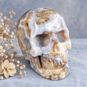 May include: A carved stone skull sculpture featuring a polished surface with a marbled pattern of translucent light blue, milky white, and earthy tan brown inclusions throughout the entire structure, showcasing natural mineral variations.