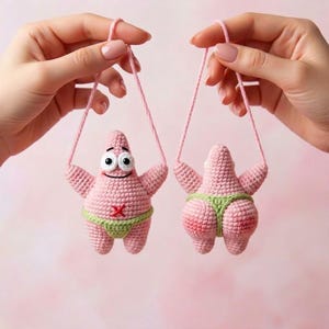 May include: Two pink crocheted starfish figures, one showing the front with a smiling face and a red X on the chest, and one showing the back. Both figures wear a light green crocheted thong and are suspended by pink yarn loops for hanging.
