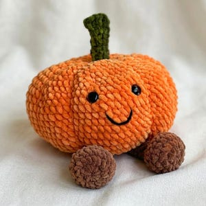 May include: A plush, crocheted pumpkin toy with a bright orange, textured body, a dark green stem, and two small brown feet. The pumpkin features two black plastic safety eyes and a stitched black smile, creating a friendly, rounded appearance.
