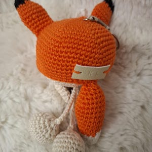 Op de afbeelding: Een gehaakte vossen-sleutelhanger met een feloranje kop en puntige oren met zwarte uiteinden. Aan de voorzijde is een klein cr&egrave;mekleurig leren label met de in reli&euml;f gedrukte letters KL bevestigd. Twee cr&egrave;mekleurige gehaakte bollen hangen eronder.