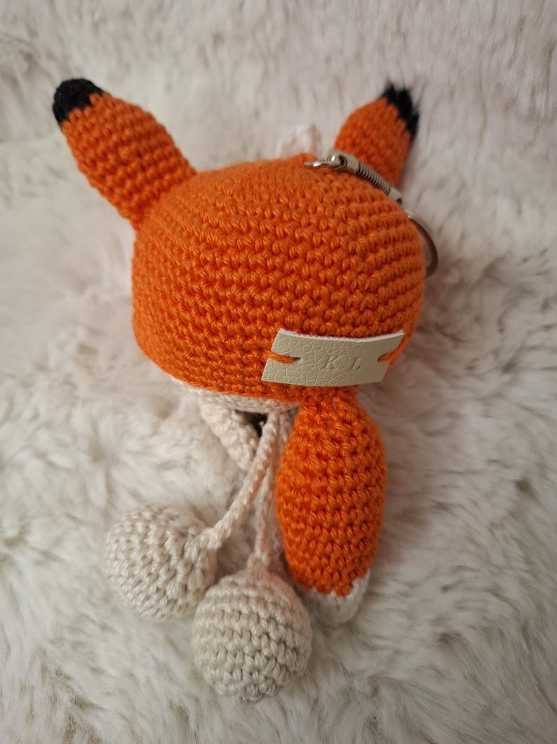 Op de afbeelding: Een gehaakte vossen-sleutelhanger met een feloranje kop en puntige oren met zwarte uiteinden. Aan de voorzijde is een klein cr&egrave;mekleurig leren label met de in reli&euml;f gedrukte letters KL bevestigd. Twee cr&egrave;mekleurige gehaakte bollen hangen eronder.