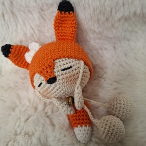 Op de afbeelding: Een gehaakte vossenpop met een feloranje capuchon met zwartgepuntte oren en een kleine witte strik. De pop heeft een cr&egrave;mekleurig gezicht met gesloten ogen, een zwarte neus en een klein bruin belletje bij de nek, met bungelende cr&egrave;mekleurige pompons.
