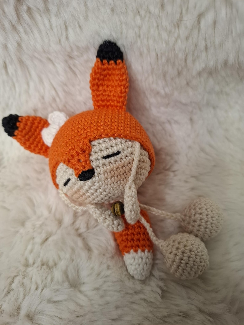 Op de afbeelding: Een gehaakte vossenpop met een feloranje capuchon met zwartgepuntte oren en een kleine witte strik. De pop heeft een cr&egrave;mekleurig gezicht met gesloten ogen, een zwarte neus en een klein bruin belletje bij de nek, met bungelende cr&egrave;mekleurige pompons.