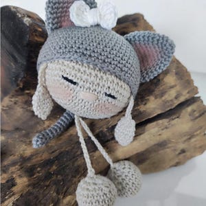 Könnte beinhalten: Eine handgefertigte Häkelpuppe in Form eines Kopfes mit grauer Katzenohren-Kapuze und einer kleinen weißen Schleife. Die Puppe hat ein beiges Gesicht mit geschlossenen Augen, rosa Wangen und einer kleinen Nase sowie zwei langen Häkelschnüren.