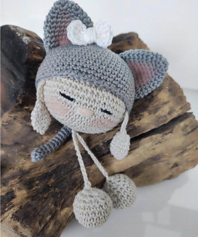 Puede incluir: Una cabeza de mu&ntilde;eca tejida a ganchillo a mano con una capucha gris de orejas de gato y un peque&ntilde;o lazo blanco. La mu&ntilde;eca tiene una cara beige con ojos cerrados, mejillas rosadas y una nariz peque&ntilde;a. Dos cuerdas finas terminan en bolas beige.