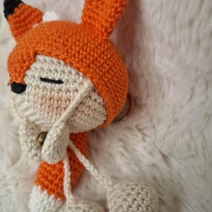 Op de afbeelding: Een handgemaakte gehaakte vossensleutelhanger met een cr&egrave;mekleurig gezicht met gesloten ogen, een zwarte neus en een oranje capuchon met puntige oren met zwarte uiteinden. De vos heeft bungelende cr&egrave;mekleurige garenkoorden met kleine bolletjes.
