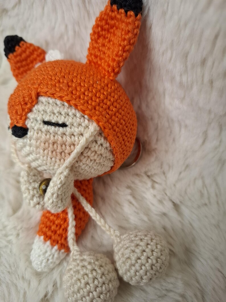 Op de afbeelding: Een handgemaakte gehaakte vossensleutelhanger met een cr&egrave;mekleurig gezicht met gesloten ogen, een zwarte neus en een oranje capuchon met puntige oren met zwarte uiteinden. De vos heeft bungelende cr&egrave;mekleurige garenkoorden met kleine bolletjes.