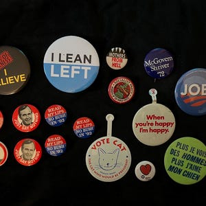 May include: A collection of vintage political campaign buttons featuring various slogans and images, including "I Lean Left", "Read My Lips No Bush in '92", "Jobs", "McGovern Shriver '72", and "Vote Cat".