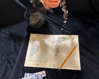 May include: An open, cream-colored stationery set with floral and scroll designs, a pencil, and several vintage stamps are displayed on a black surface. A vase with a peach-colored rose and dried flowers is in the background.