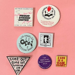 以下が含まれることがあります： 様々なメッセージとデザインが施された、ヴィンテージのボタンピン8個のセット。ピンはほとんどが白地に黒の文字とグラフィック。一部のピンには虹のデザインが施されています。ピンはピンク色の背景の上に置かれています。