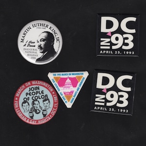 May include: Four round buttons with different designs. The top left button is black and white with a portrait of Martin Luther King Jr. and the text "Martin Luther King Jr. (1929-1968) I Have A Dream Inaugural National Holiday 1986". The top right button is black with white text that says "DC ≥93 APRIL 25, 1993". The bottom left button is red with white text that says "*MARCH ON WASHINGTON JOIN PEOPLE OF COLOR FOR LESBIAN & GAY RIGHTS 11, 1987*" and a picture of three men. The bottom right button is black with white text that says "DC ≥93 APRIL 25, 1993".