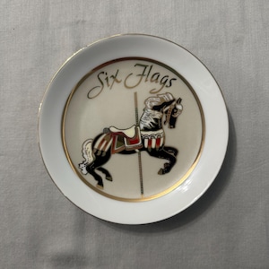 May include: A round white ceramic plate with a gold rim featuring a central illustration of a black carousel horse with a red and white saddle. The text Six Flags is printed in gold script above the horse, which is mounted on a vertical pole.