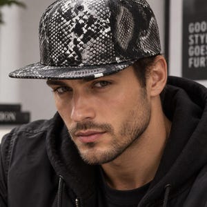 Puede incluir: Una gorra snapback con un patrón de piel de serpiente en blanco y negro de alto contraste. La gorra tiene una corona estructurada, una visera plana y un pequeño botón en la parte superior central. El material muestra una textura de escamas detallada.