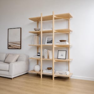 May include: A tall, light-coloured wooden bookshelf featuring a vertical support structure that divides the shelves into sections. The unit holds various books, small decorative bowls, and framed art pieces arranged across multiple open levels.