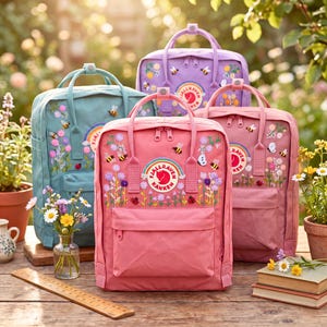 Op de afbeelding: Vier Fjallraven Kanken rugzakken in lichtblauw, lavendel, roze en oudroze, elk voorzien van geborduurde wilde bloemen, bijen en lieveheersbeestjes. Elke tas heeft een rond logopatch met een regenboog, twee handvatten en een voorvak met rits.