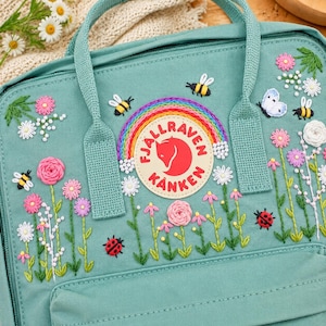 Könnte beinhalten: Ein mintgrüner Rucksack mit einer Stickerei aus Regenbogen, Bienen, Marienkäfern, einem Schmetterling und verschiedenen bunten Blumen. In der Mitte befindet sich ein kreisförmiger Fjällräven Kånken-Aufnäher mit einem roten Fuchssymbol.