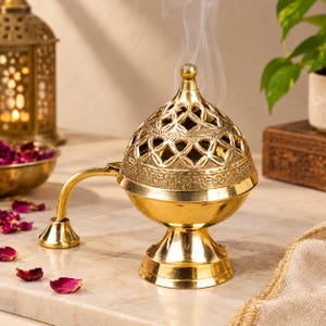 May include: A polished gold-toned metal incense burner with a dome-shaped lid featuring intricate cut-out patterns and a textured band. The burner has a flared base and a curved handle extending to the side, with thin wisps of smoke rising upwards.