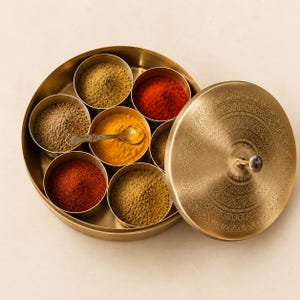 Puede incluir: Un especiero redondo de latón con una tapa a juego que presenta intrincados patrones grabados. El recipiente contiene siete cuencos pequeños extraíbles llenos de diversas especias molidas de colores, incluyendo polvos rojos, amarillos y marrones, y una pequeña cuchara de metal.