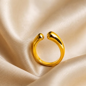 May include: A polished gold-tone open ring featuring a smooth, rounded, and fluid design. The band is crafted with a continuous, thick, and curved silhouette that tapers slightly at the two ends, creating a gap at the top of the circular shape.