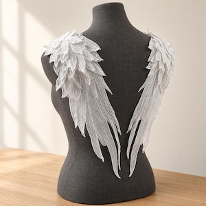 Puede incluir: Un par de alas de ángel de encaje blanco en aplique con detalles de bordado intrincados que parecen plumas. La tela delicada y semitransparente tiene forma de segmentos de ala superpuestos para añadir a prendas textiles.