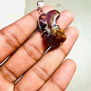 May include: A polished, irregular-shaped fire agate pendant set in a silver-toned metal wire frame. The gemstone displays deep reddish-brown and amber hues with translucent, swirling internal patterns and a metallic luster throughout the stone.