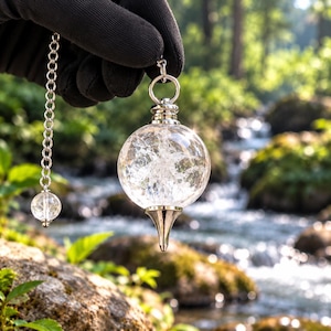 May include: A clear, spherical crackle quartz pendulum featuring a silver-toned metal cap and a pointed base. The pendulum hangs from a silver-toned metal chain that includes a smaller, matching spherical crackle quartz bead near the top.